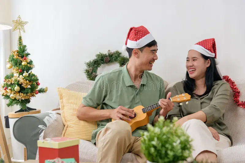 A happy couple sits on the living room floor in front of a decorated Christmas tree, holding a jar of lights as they share happy Christmas and New Year wishes along with Christian wedding anniversary wishes for husband.